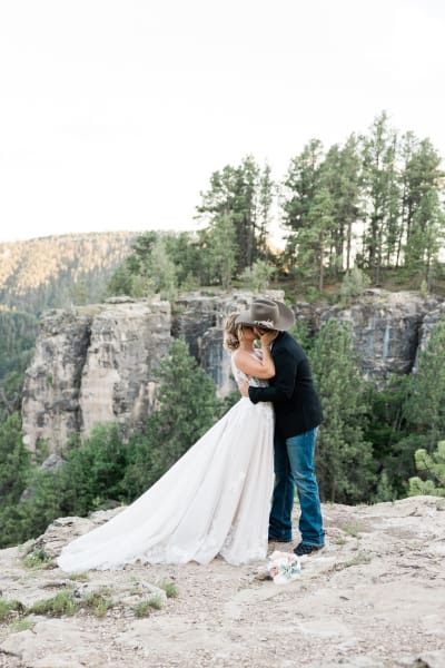 A bride and groom embrace and kiss standing on a vista. The bride is dressed in white and the groom wears a cowboy hat.