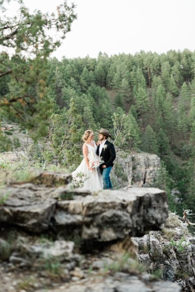 A bride and groom stand looking at each other among rocks and evergreen trees, looking at each other lovingly.