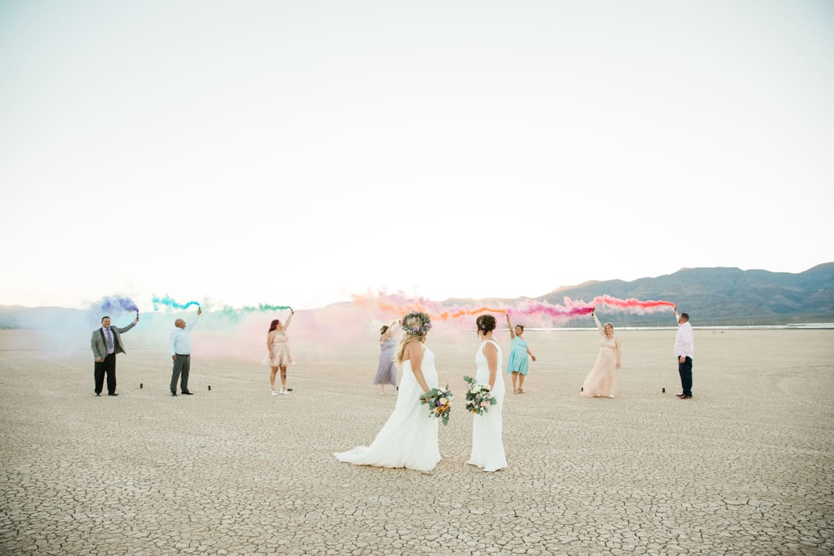 Brides watching their wedding guests hold colored smoke bombs in the desert.