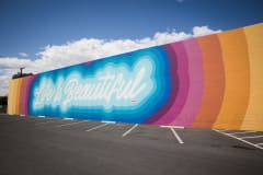 The words \"life is beautiful\" are written in white script and then gradiants of blues, pinks, and oranges surround the lettering to cover the entire side of a building.