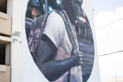 A black man wearing a cowboy hat and white t-shirt, carrying a backpack, looks at the viewer, like an artistic rendering of a photograph. The artwork is inside a large circle on the side of a white building.