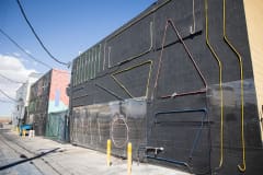 Multiple colors of thin neon lights are bent into geometric shapes and attached to the side of a building's black wall. They aren't lit up because it's day time.