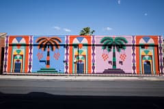 A photo of the side of a building and blue skies. The building is painted in a variety of colors to depict arched doorways and giant palm trees.