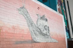 A brick wall mural featuring a black and white snail carrying a smoking skull as a shell on its back with three black birds nestled right in the cranium. In the white, pink, and red background, you’ll see a desert scape in shades of orange, red and pink, with the distant silhouette of palm trees, desert plants and mountains.