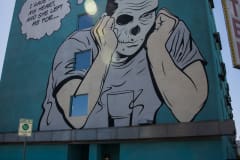 A mural of a retro-comic styled man with a skeletal face is sitting with a drink with a thought bubble coming from his head that says, “I gave her my heart, she left me for…”