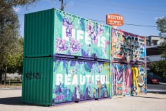 Two large cargo containers are stacked and tagged with grafitti. They read \"Life Is Beautiful\" in white letters on a mint green background and surrounded by purple flowers.