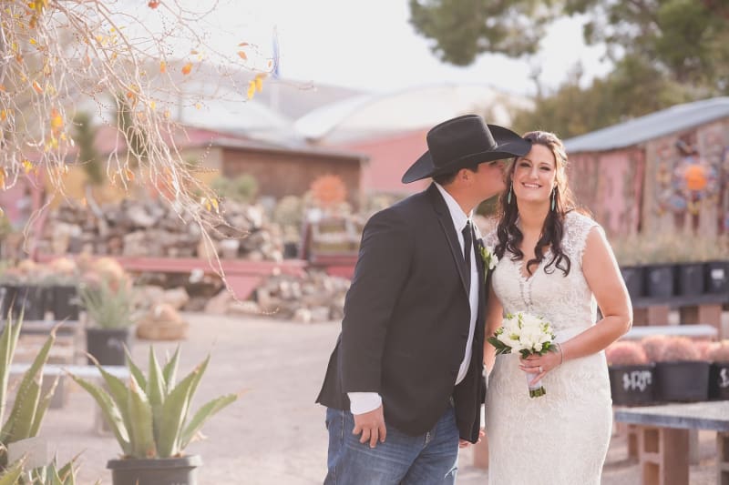 A groom in a cowboy hat leans in to kiss his smiling bride as they pose for wedding photos at Cactus Joe’s Blue Diamond Nursery .