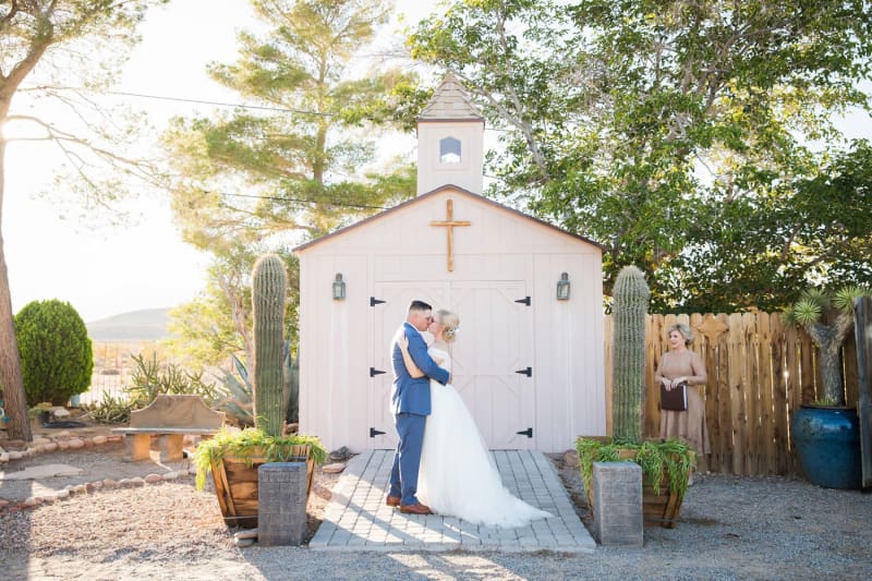 A wide shot of a groom and bride kissing in front of a small wooden chapel flanked by two tall cacti.