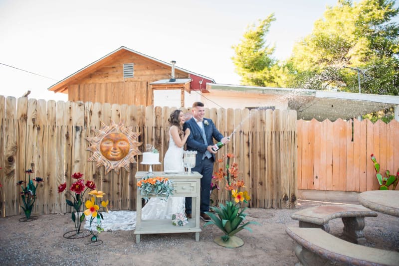 A just-married couple sprays Champagne from behind a mini celebration table as they celebrate amongst metal plant sculptures.
