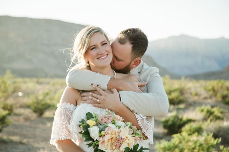 A medium shot of a bride and groom embracing in the desert with mountains behind them.