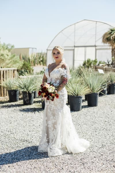 A full body portrait of a bride in front of a greenhouse with cacti on the ground just behind her.