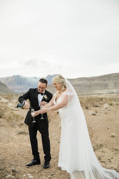 A groom and bride pour themselves glasses of Champagne in the desert with mountains in the background.