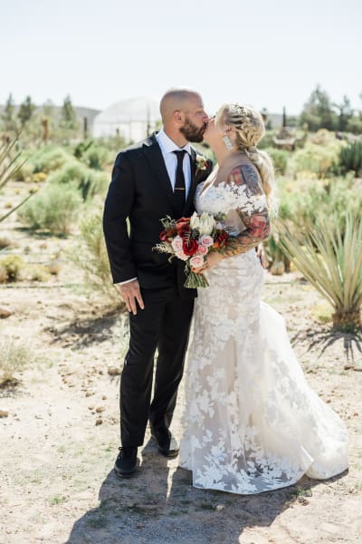 A bride and groom kiss in a desert landscape full of cacti and bushes.