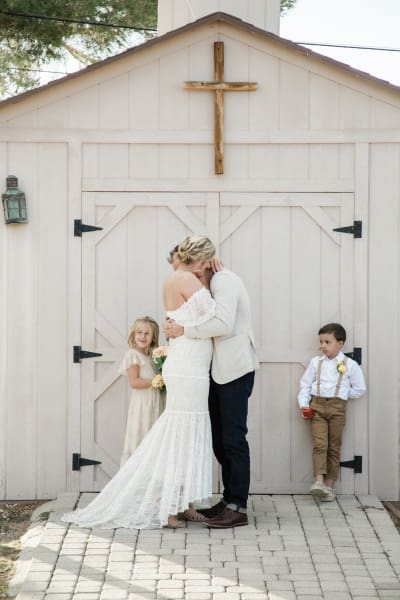 A bride and groom hug while two children stand beside them in front of a small wooden chapel.