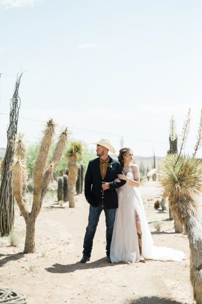 A groom and bride hold each other and look opposite ways as they stand between rows of Joshua Trees.