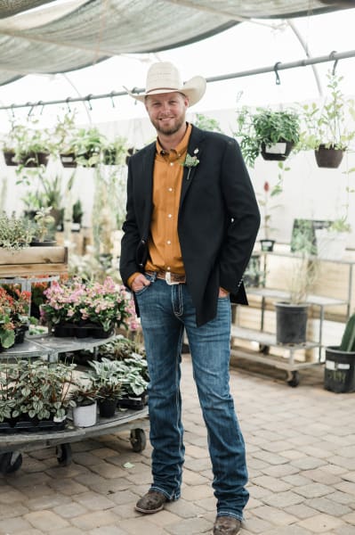 A groom in a cowboy hat, boots, jeans and a black sport coat poses amongst plants on display at Cactus Joe’s.