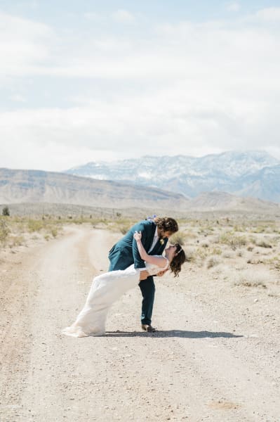 A groom dips his bride backwards on a dirt road with the snow-covered mountains of Red Rock Canyon in the background.