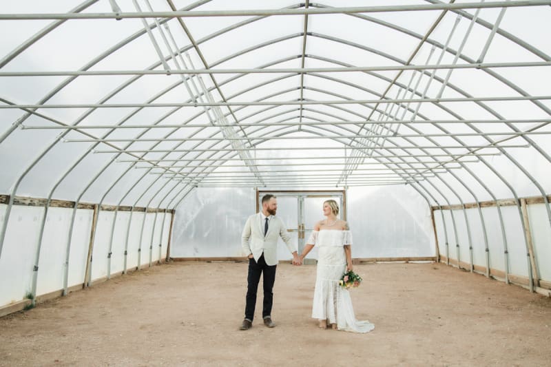 A groom and bride hold hands while standing in the center of an empty greenhouse.