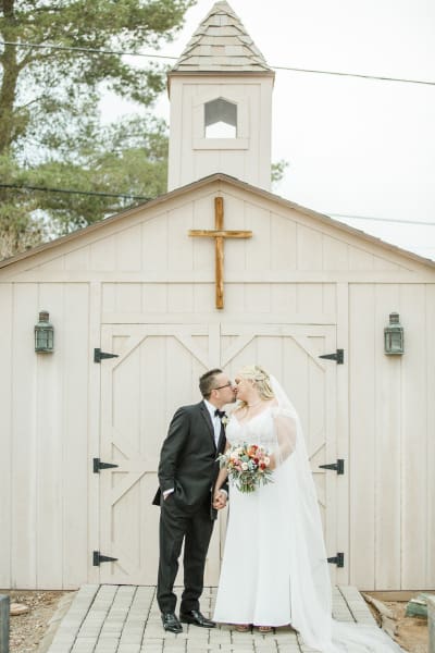 A groom and bride kiss while standing in front of the doors of a little wooden chapel.