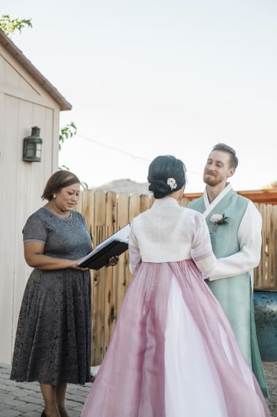 A wedding officiant performs a marriage for a couple in front of a little wooden chapel.