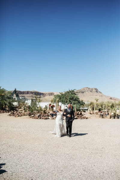 A wide shot of a groom escorting his new bride towards the camera with desert mountains in the background.