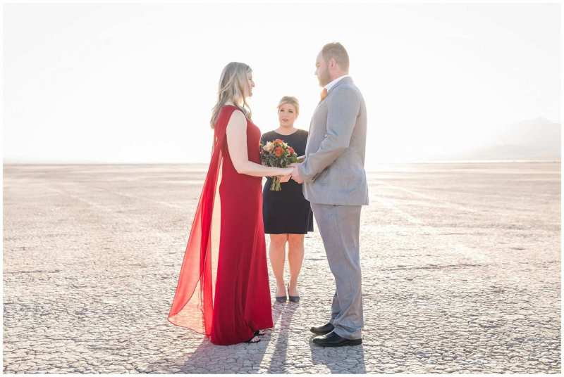 A woman in a long flowy red dress faces her fiance as they stand together with a wedding officiant. Their wedding ceremony is taking place on the Eldorado Dry Lake Bed. Bright sun fills the image from the rear.
