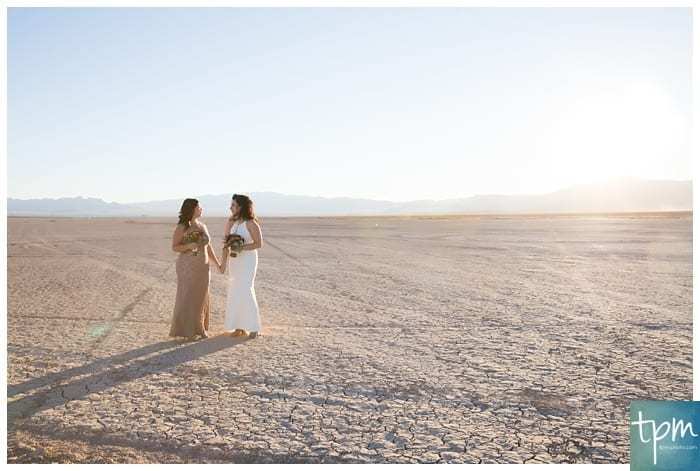 A wide shot of two brides standing together and holding hands. They cast long shadows on the parched ground of the Dry Lake Bed near Las Vegas.
