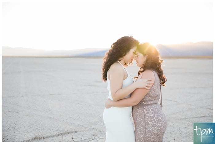 Two women place their foreheads together and stand holding each other on the Dry Lake Bed south of Boulder City near Las Vegas, Nevada. They are both wearing elegant sleeveless dresses. The sunsets over the mountains behind them.
