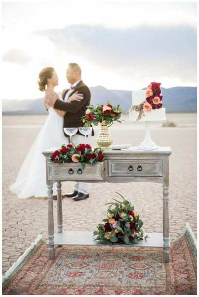 A mini celebration table is seen with flowers, a wedding cake and Champagne glasses. Behind the table a bride and groom are out of focus against the setting sun. The stylish table sits on a red decorative rug that has been placed on the ground at the Dry Lake Bed in Southern Nevada.