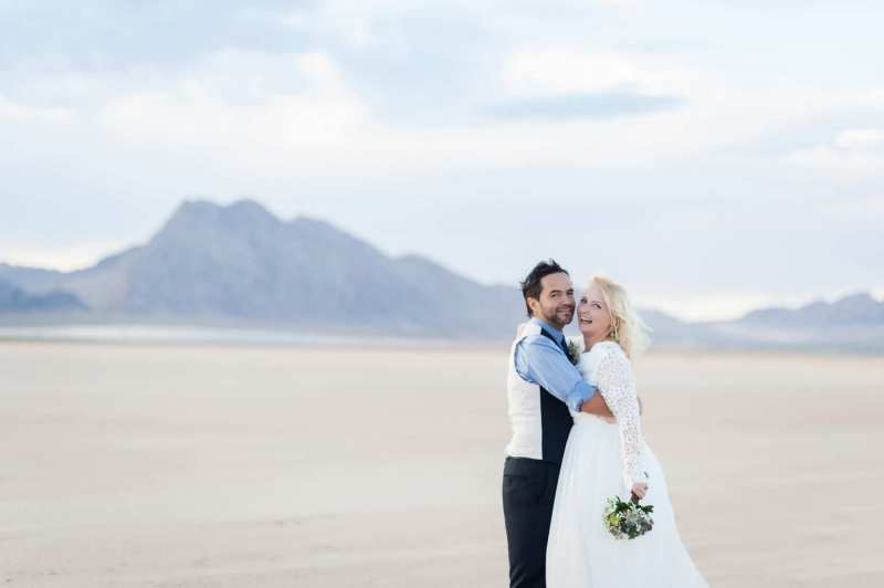 A groom and bride hold each other closely as they turn towards the camera and laugh. They are posing for photos following their wedding ceremony at the Eldorado Dry Lake Bed outside of Las Vegas.