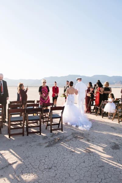 A man in a white military uniform escorts a bride down the aisle. A wedding ceremony is being held on the Eldorado Dry Lake Bed outside of Las Vegas, Nevada. Guests have stood up to their chairs to watch the bride walk towards the front of the gathering.