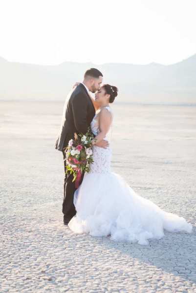 A medium shot of a groom and his bride embracing on their wedding day. They have just gotten married on the Dry Lake Bed near Boulder City and South of Las Vegas, Nevada. The ground they stand on is dry and cracked in an artful way.