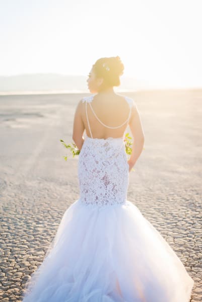 A bride in a white wedding dress faces away from the camera and into the sun so that the photograph captures her bubbly dress as it is contrasted against the stark landscape of the Dry Lake Bed.