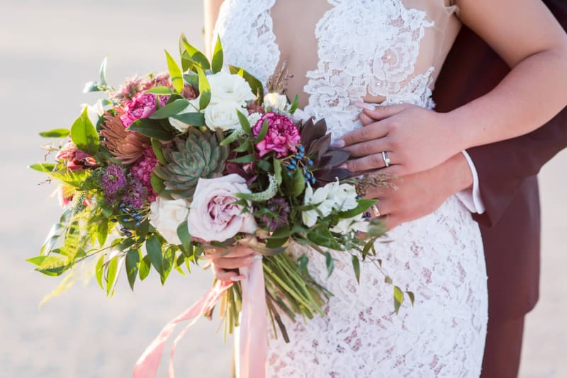 A close up photo of a bridal bouquet of flowers which are held in the right hand of the bride while her left hand is placed against her body where it meets her new husband's hand. 