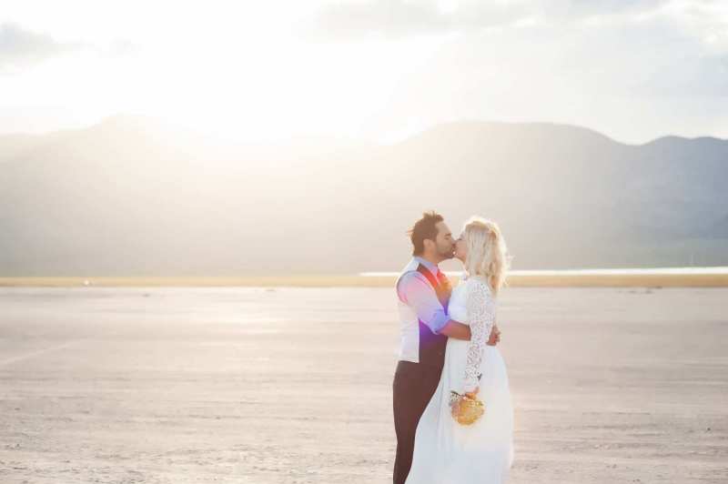 Bright late afternoon sun illuminates a kissing husband and wife. They are celebrating their wedding day on the Dry Lake Bed south of Boulder City, Nevada.
