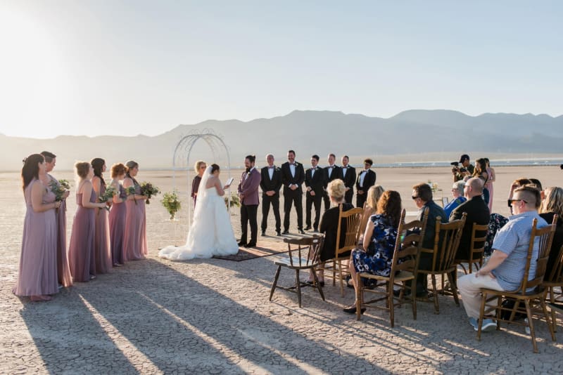 A wedding is being held at the Dry Lake Bed in Las Vegas. At the center is the bride and groom and the officiant. They are flanked by their bridesmaids and groomsmen. Several guests sit in chairs arranged in front of them.
