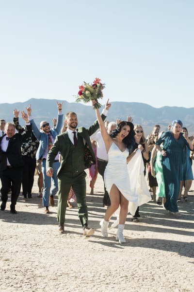 A bride and groom and their wedding party all walk towards the camera. The entire group is raising their arms, pumping their fists and hooting and hollering with joy. They are on the Dry Lake Bed near Las Vegas.