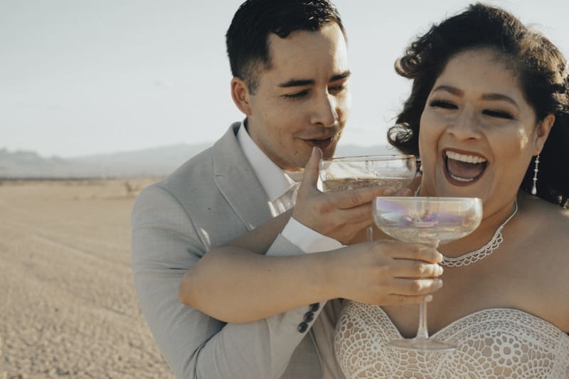 A bride and groom lock arms and drink Champagne from coupe glasses. They are toasting their marriage following their wedding on the Eldorado Dry Lake Bed outside of Boulder City and Las Vegas, Nevada.