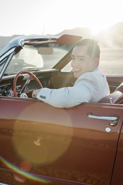 A man in a light tan suit rests his arm on the top of the door of a red Ford Mustang Convertible. He is sitting in the driver's seat and smiling as the sunlight hits him from behind.