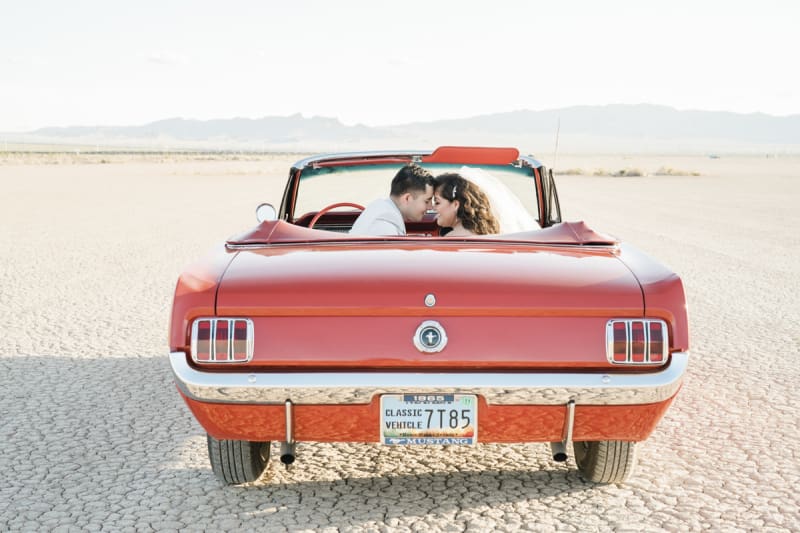 A man and woman sit in the front seats of a 1965 Ford Mustang convertible. The photo is taken from behind the car. The top is down. They are posing for wedding photos on the Dry Lake Bed south of Las Vegas, Nevada.