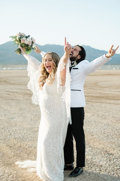 A couple celebrates their wedding on the Dry Lake Bed in Boulder City, Nevada. They stand together with the man behind the woman and each raise their arms and scream with excitement.