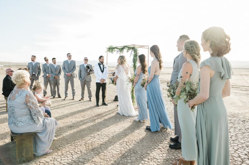 A wedding is being held on the Eldorado Dry Lake Bed. The bride and groom are in the middle and are flanked by their wedding party. Guests are seen sitting on wooden benches.