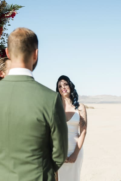 A bride stands with her groom. The photo is taken from behind the groom with the bride facing the camera and smiling at the groom. They are getting married on the Eldorado Dry Lake Bed.