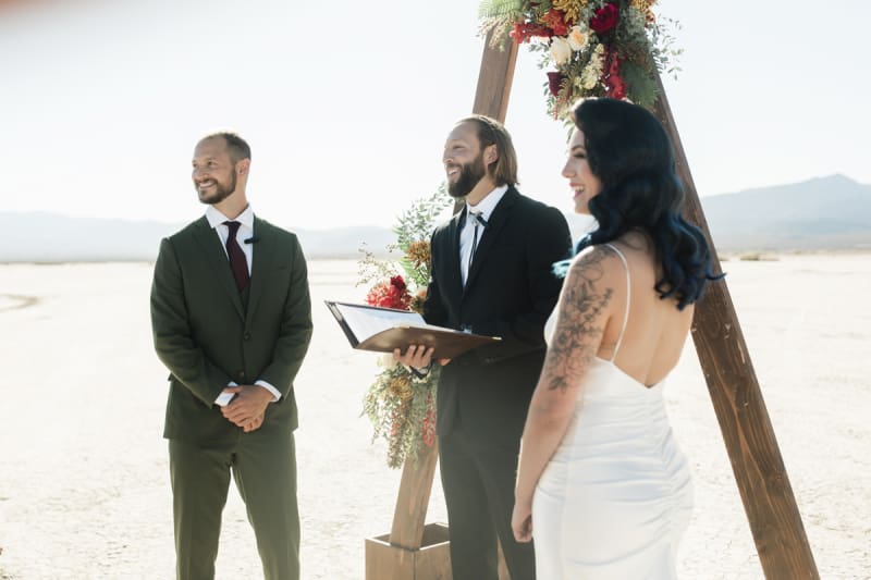 On the Eldorado Dry Lake Bed, a groom and bride turn towards the left of the picture to look off camera as their wedding officiant addresses their guests. There is a wooden triangular shaped arbor decorated with flowers.