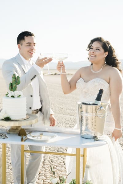 A groom and a bride in a strapless white wedding dress toast each other with Champagne coupe glasses. A wedding cake and an ice bucket with a bottle sit on a table which they stand behind. They are celebrating their wedding on the Dry Lake Bed.