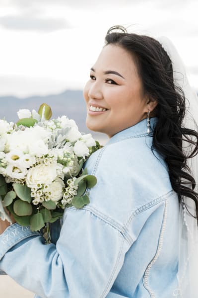 A close up of a bride posing for wedding photos. She is holding her flowers about shoulder high as she turns her head back to the left and towards the camera. She is wearing a light blue denim jacket and a white veil.