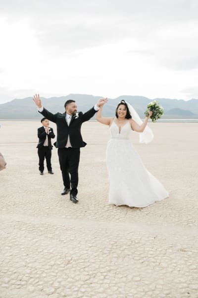 A just married couple raises their hands in the air to celebrate their wedding. They walk on the dry cracked ground of the Dry Lake Bed, while a young boy claps for them in the background.