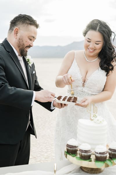 A groom and bride use golden forks to dig into a piece of chocolate wedding cake. The cake is being served at their wedding on the Dry Lake Bed near Las Vegas.