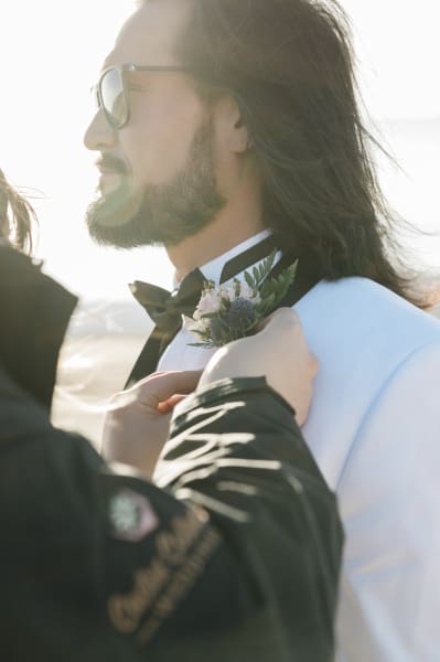 A boutonniere is pinned onto a groom's lapel. He is wearing a white tuxedo jacket and a black bowtie.He sports a beard, long hair and sunglasses.