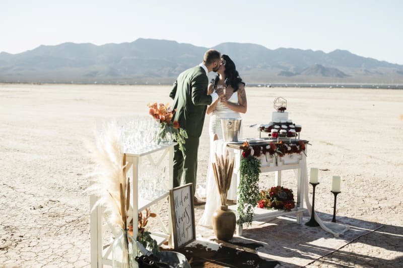 A groom and bride kiss as they celebrate their wedding with cupcakes and Champagne. They are standing behind a pair of small tables arranged with the treats, flowers candles and a sign that reads, "All of me loves all of you".